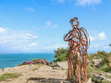 A metal sculpture of two people embracing on a rocky cliff with the ocean and blue sky in the background at Watlands in Mynytho near Abersoch