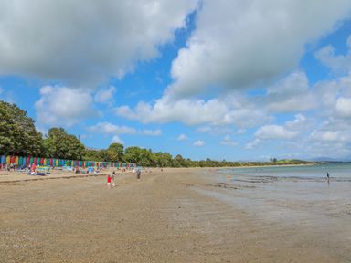 A beach with people playing and colorful beach huts near trees and water at Watlands in Mynytho near Abersoch