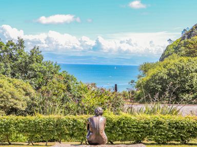 A bronze statue of a person sitting on a stone with a view of the sea and trees at Watlands in Mynytho near Abersoch