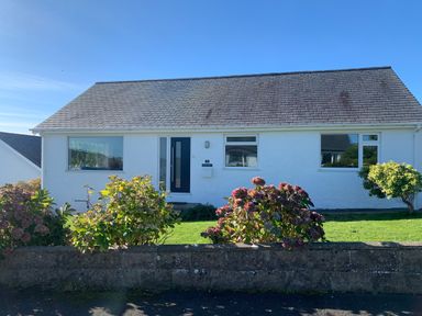 A single-story white house with a dark door and three windows a garden with bushes and flowers in front at Watlands Mynytho near Abersoch
