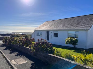 A white house with a slate roof and a garden with bushes and a small tree overlooking a coastal landscape at Watlands in Mynytho near Abersoch