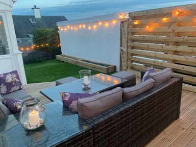 An outdoor seating area with rattan furniture cushions and candles on glass tables beside a wooden fence and lawn at Watlands in Mynytho near Abersoch