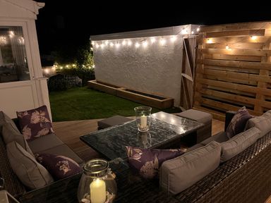 An outdoor seating area with cushioned sofas around a glass table with candles and string lights on walls and wooden panels at night at Watlands in Mynytho near Abersoch