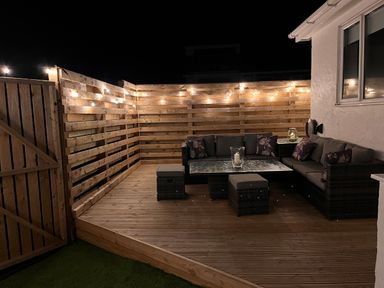 A wooden fenced patio with string lights a sectional sofa table and stools at Watlands in Mynytho near Abersoch