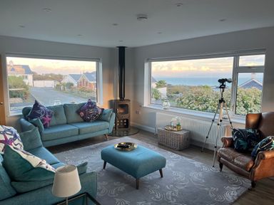 A living room with blue sofas and an armchair with large windows showing a sea view at Watlands in Mynytho near Abersoch