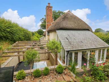 An outdoor area with a wooden pergola and hot tub next to a thatched cottage with a slate roof at Rose Cottage in Okehampton
