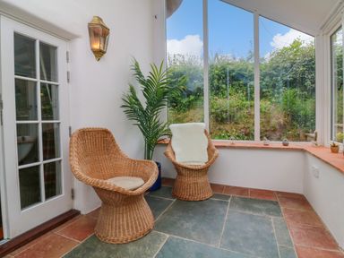 A sunroom with two wicker chairs a potted plant a wall-mounted light and large windows showing a garden at Rose Cottage in Okehampton