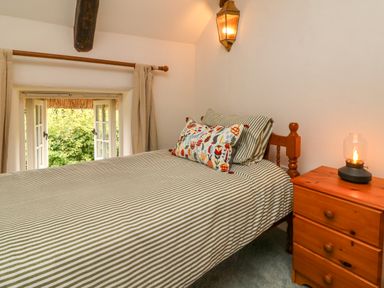 A bedroom with a bed with striped bedding a wooden nightstand with a lamp and an open window at Rose Cottage in Okehampton