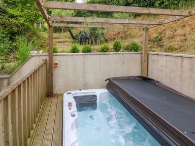 An outdoor hot tub with half cover on a wooden deck under a wooden pergola with string lights and a patio dining set on grass behind a concrete wall at Rose Cottage in Okehampton