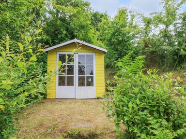 A yellow garden shed with white double doors surrounded by green bushes and trees at Rose Cottage in Okehampton