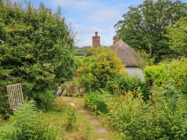 A garden path with stone slabs leading to a thatched roof cottage surrounded by bushes and trees