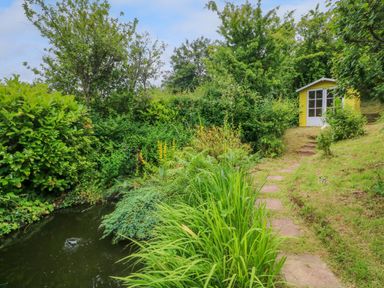 A garden with a pond a stone path leading to a yellow shed surrounded by trees and bushes at Rose Cottage in Okehampton