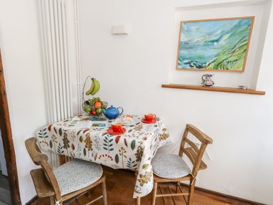 A small dining area with a table covered by a floral tablecloth two wooden chairs with patterned cushions a fruit basket with bananas grapes and oranges a blue teapot two red cups with saucers and a pastry on a plate a framed landscape painting and decorative items on a wall shelf at Tiddlers Cottage in Uploders near Bridport