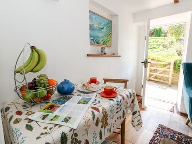 A small dining area with a table covered with a patterned tablecloth holding fruit, a blue teapot, two orange cups and saucers with pastries and an open magazine with a door open to a garden at Tiddlers Cottage in Uploders near Bridport