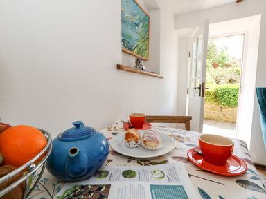 A kitchen table with a blue teapot orange cups pastries a fruit bowl and an open door showing a garden at Tiddlers Cottage in Uploders near Bridport