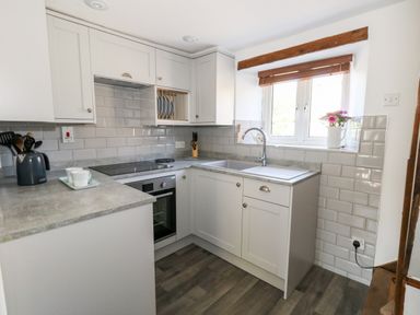 A kitchen with white cabinets a countertop with a kettle and cups a stove an oven a sink under a window and flowers in a vase at Tiddlers Cottage in Uploders near Bridport