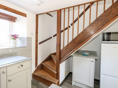 A small kitchen area with wooden stairs a window with flowers on the sill a refrigerator a microwave and white cabinets at Tiddlers Cottage in Uploders near Bridport