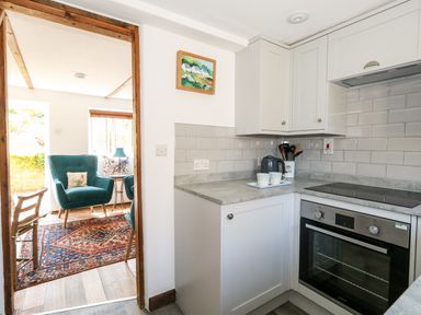 A kitchen with white cabinets grey countertop electric stove and oven looking into a living area with blue armchairs and a patterned rug at Tiddlers Cottage Uploders near Bridport