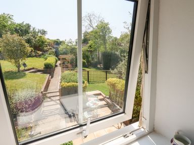 View of a garden with trees and bushes from a window looking out onto a patio area with a table and chairs at Tiddlers Cottage in Uploders near Bridport