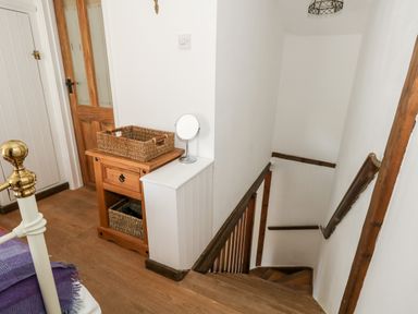 A staircase with wooden handrails next to a small wooden cabinet with two wicker baskets and a round mirror at Tiddlers Cottage in Uploders near Bridport