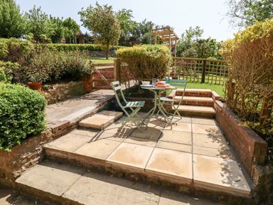 An outdoor patio area with two chairs and a small round table set with food surrounded by hedges and steps at Tiddlers Cottage in Uploders near Bridport