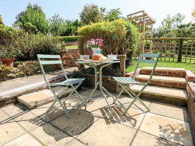 A garden patio with two metal chairs and a small table with bread and flowers at Tiddlers Cottage in Uploders near Bridport