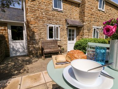 A patio table with bread bowls glasses and flowers outside a stone cottage with a wooden bench at Tiddlers Cottage in Uploders near Bridport