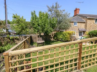 A garden with wooden fence tables and chairs trees and a stone house in the background at Tiddlers Cottage in Uploders near Bridport