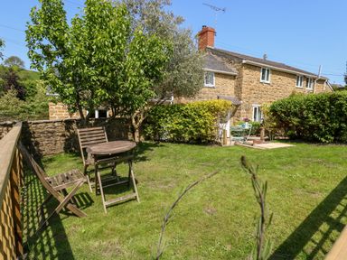 A garden with wooden chairs and a table near a stone house with trees and hedges at Tiddlers Cottage in Uploders near Bridport