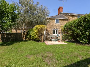 A stone cottage with two windows and a chimney in a garden with a table and chairs on a small patio at Tiddlers Cottage Uploders near Bridport