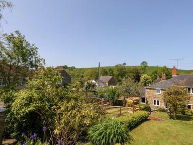 A garden with bushes and flowers overlooking stone houses and green hills at Tiddlers Cottage in Uploders near Bridport