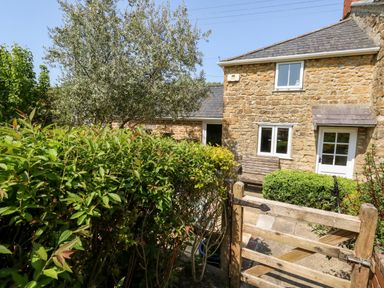 A stone house with a wooden gate and green bushes in front at Tiddlers Cottage in Uploders near Bridport