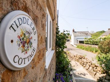 A close up of a sign reading Tiddlers Cottage on a stone wall with a road and houses in the background at Tiddlers Cottage Uploders near Bridport