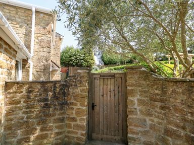 A wooden gate labeled Tiddlers Cottage set in a stone wall with trees and part of a stone building nearby at Tiddlers Cottage in Uploders near Bridport