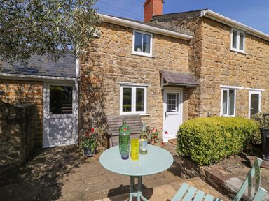 A stone cottage exterior with a small patio table and chairs and a wooden bench at Tiddlers Cottage in Uploders near Bridport
