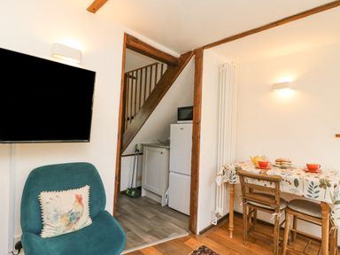 A room with a green chair with a rooster cushion a wall-mounted tv a small dining table with two chairs and a kitchenette under stairs at Tiddlers Cottage in Uploders near Bridport