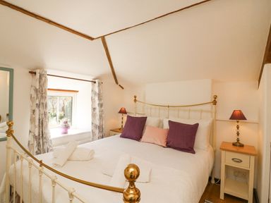 A bedroom with a bed with white bedding and purple pillows a window with floral curtains and two bedside tables with lamps at Tiddlers Cottage Uploders near Bridport