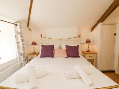 A bedroom with a double bed with purple and pink pillows two bedside tables with lamps and rolled white towels at Tiddlers Cottage in Uploders near Bridport