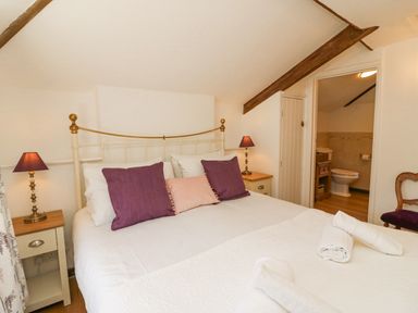 A bedroom with a double bed with purple and pink pillows bedside tables with lamps and a bathroom visible through an open door at Tiddlers Cottage Uploders near Bridport