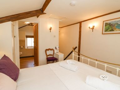 A bedroom with a white metal bed frame and rolled towels on the bed a wooden chair with a purple cushion a small white side table a doorway leading to a bathroom a framed painting on the wall at Tiddlers Cottage in Uploders near Bridport