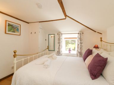 A bedroom with a white bed with purple pillows a window with floral curtains and a framed picture on the wall at Tiddlers Cottage Uploders near Bridport