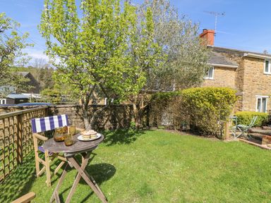 A small garden with a wooden table and chair with drinks and cakes near a stone house at Tiddlers Cottage in Uploders near Bridport