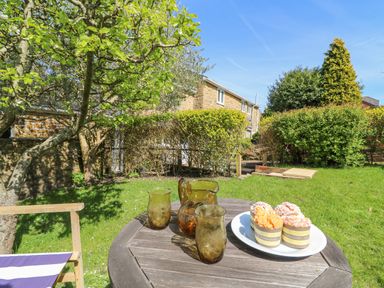 A garden with a wooden table holding three amber glasses a pitcher and a plate of cupcakes at Tiddlers Cottage in Uploders near Bridport