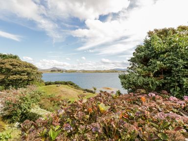 A garden with flowers and trees overlooking a body of water with hills in the distance at Sea Watch House in Roundstone County Galway