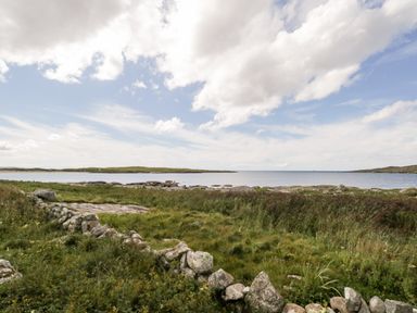 A coastal landscape with grass fields stones and calm water under a partly cloudy sky at Sea Watch House in Roundstone County Galway