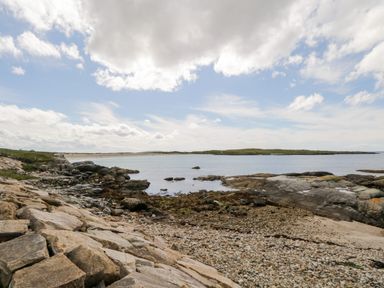 A rocky shoreline with a pebble beach and calm water under a partly cloudy sky at Sea Watch House in Roundstone County Galway