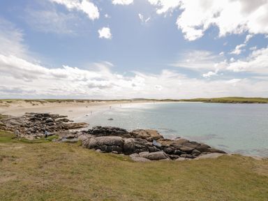 A beach with sand rocks and grass next to clear water under a cloudy sky at Sea Watch House in Roundstone County Galway