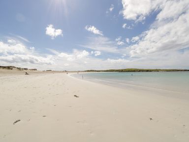 A sandy beach with footprints and a few people near the water and in the water under a partly cloudy sky at Sea Watch House in Roundstone County Galway