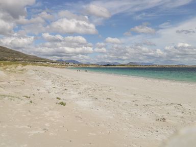 A sandy beach with people walking near the shoreline with mountains and a partly cloudy sky in the background at Sea Watch House in Roundstone County Galway