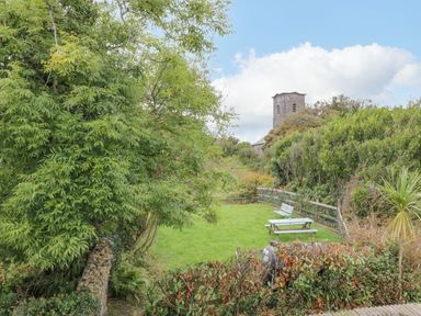 A garden with benches and picnic tables surrounded by trees and bushes with a stone building tower in the background at Sea Watch House in Roundstone County Galway